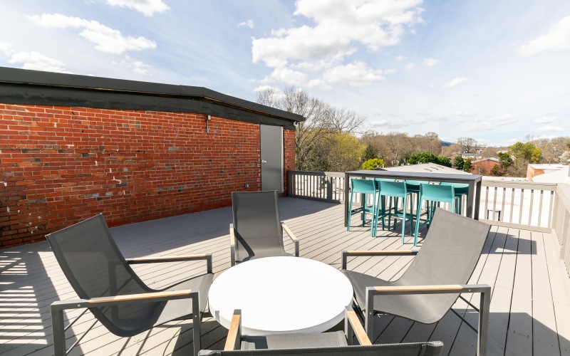 A rooftop patio featuring four modern outdoor chairs around a white circular table in the foreground. In the background, there is a high bar table with stools. The patio has gray decking, a brick wall on one side, and overlooks a view of trees and houses.