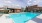 An outdoor pool area with lounge chairs and tables with umbrellas. A brick building is next to the pool, featuring restrooms and a shaded seating area. Trees and a red brick building surround the area. The pool has depth markers reading "3 FT" and "4 FT.