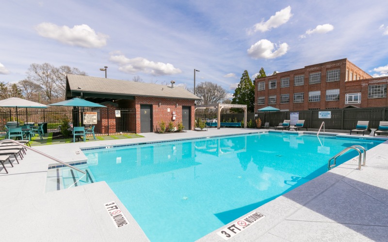 An outdoor pool area with lounge chairs and tables with umbrellas. A brick building is next to the pool, featuring restrooms and a shaded seating area. Trees and a red brick building surround the area. The pool has depth markers reading "3 FT" and "4 FT.