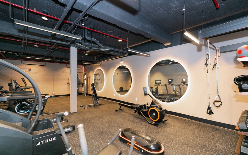 A modern gym with various exercise equipment, including treadmills, stationary bikes, and weight machines. The room features circular mirrors on the walls and a sleek, industrial ceiling with exposed pipes. The lighting is bright, providing a clean and co